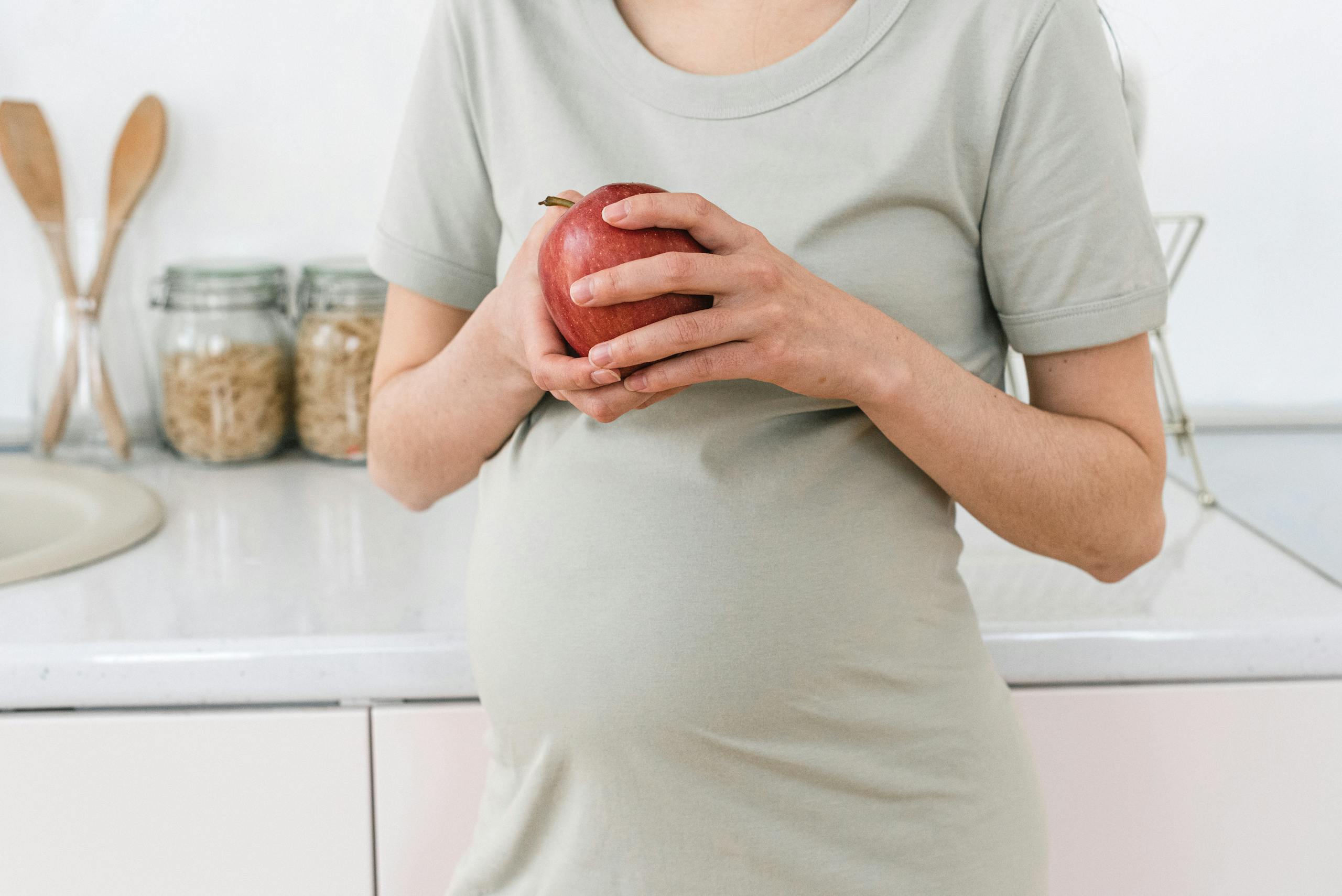 Unrecognizable pregnant female with ripe red apple for healthy diet in hands standing near counter in light kitchen at home